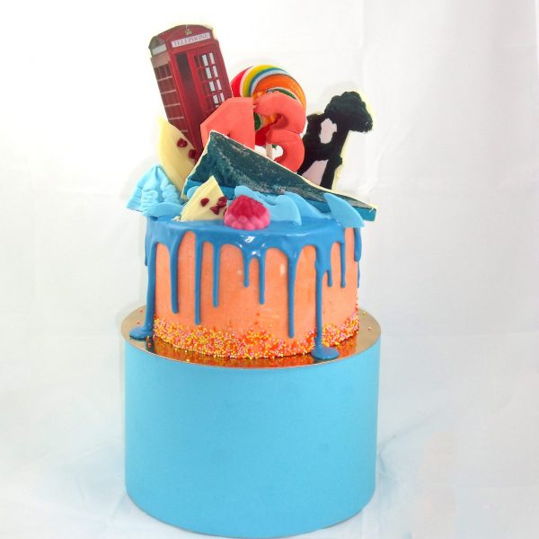 Tarta Drip Cake de dos pisos de color naranja y azul con cobertura de chocolate color azul chorreando en forma de gotas desde la parte superior y decorada con una combinacion de golosinas y diferentes modelados de fondant
