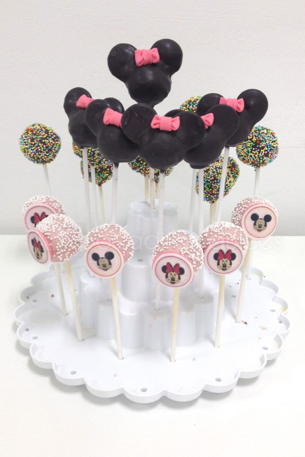 Bola de bizcocho cubierta de chocolate de colores inserado en un palo de chupa chups , unos con virutas de colores y otras con semicirculos negras de fondant a modo de orejas de Minnie Mouse y un lazo de color rosa.