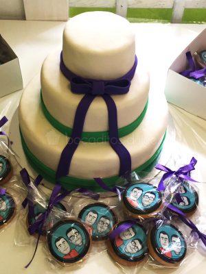 Tarta de tres pisos cubiertos de fondant blanco y decorados con cintas de fondant color verde y lazo en fondant azul, galletas de mantequilla con dibujo personalizado en papel de azúcar enbolsadas individualmente.