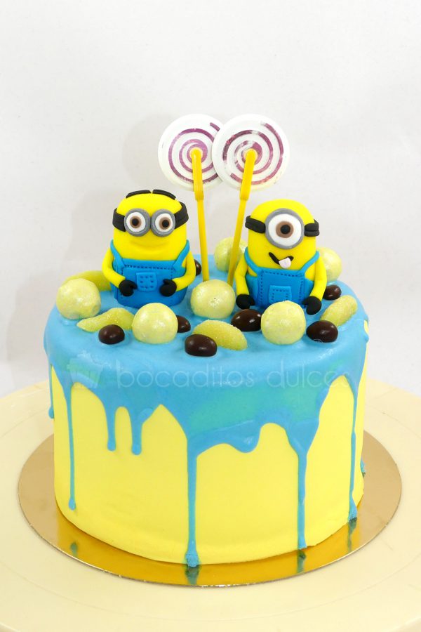 Tarta cubierta de buttercream de color amarillo con chocolate derretido por encima de color azul, decorado con chuches, y dos muñecos de la pelicula minions de fondant.