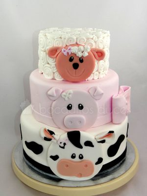 tarta de tres pisos, el primer piso forrado con fondant blanco y con pequeñas manchas negra y decorado con la cara de una vaca en fondant, segundo piso forrado de fondant rosa decorado con la cara de un cerdito y un lazo en fondant, el último piso forrado de fondant blanco decorado con pequños circulos para recrear la lana de una oveja y decorada con la cara de una oveja en fondant.