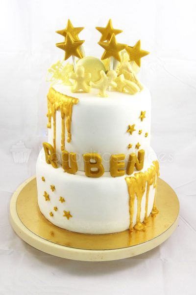 Tarta de dos pisos forrados en fondant blanco, decoracion de estrellas y nombre del niño en fondon dorado, chocolate fundido en color dorado y chocolatinas con forma de muñeco en chocolate blanco.