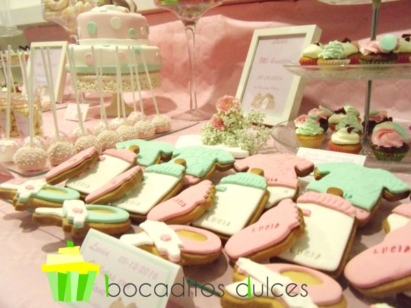 Mesa dulce en tonos rosa y verde pastel, con cake pops cubiertos de candy mels rosa y virutas de caramelo blancas, mini cupcakes decorados con buttercream rosa, verde , blanca y galletas de mantequilla con la forma de un pie de bebe, chupete y body decorados con fondant rosa y verde.