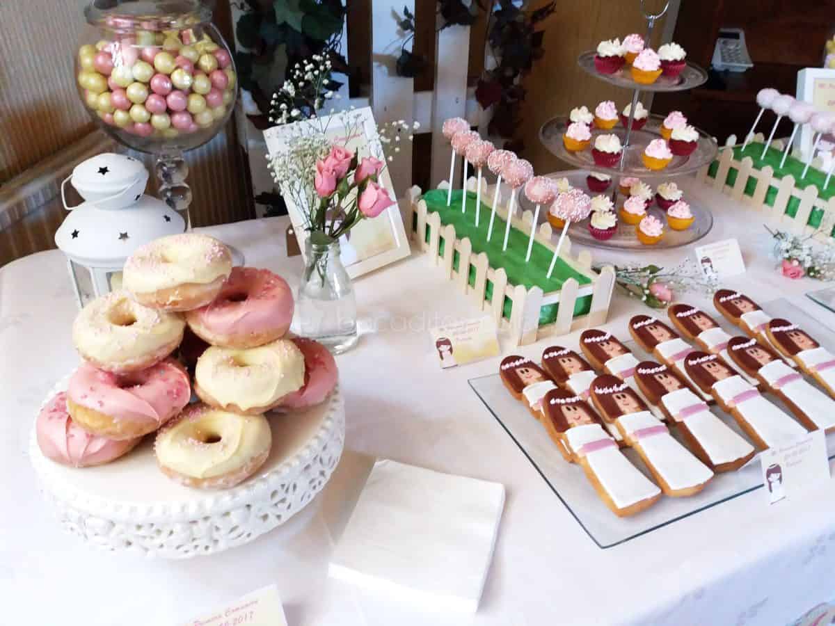 Mesa dulce compuesta de tarritos de cristal con batido de fresa, distintos tipos de golosinas, cake pops cubiertos de candy mels blanco y rosa decorados con bolitas de caramelos de color rosa y blanco, chocolatinas individiales, mini cupcakes de fresa y chocolate blanco, galletas de mantequilla con forma de niña de comunion decoradas con fondant, y donuts con graseado de color blanco y rosa.
