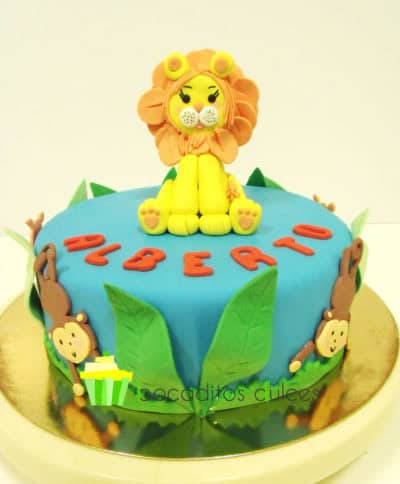 tarta cubierta en fondant azul, con monos decorativos alrededor de la tarta de fondant, hojas en fondant de color verde, el nombre de alberto en color rojo de fondant, y un pequeño leon de fondant