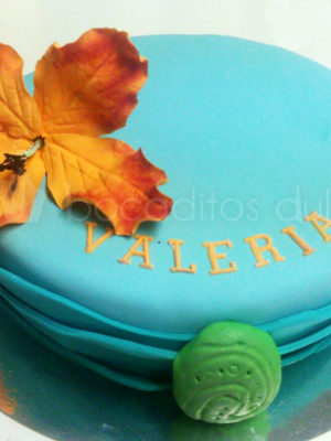 Tarta cubierta de fondant azul, con flor de color naranja de fondant nombre de Valeria en color naranja en fondant y colgante de la pelicula en fondant verde.