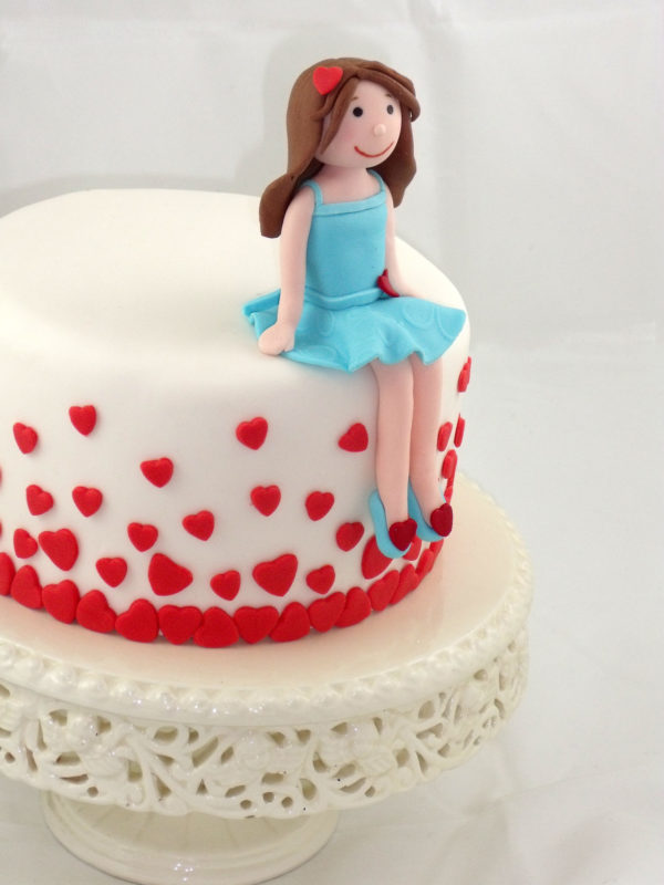 Tarta cubierta de fondant blanco con pequeños corzones de color rojo y una muñeca de fondant sentada sobre la tarta