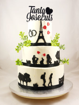 Tarta de tres pisos, cubierta de buttercream blanca, con pequeñas figuras alrededor de la tarta en negro y decorada con hojas y un letrero con el nombre de los novios