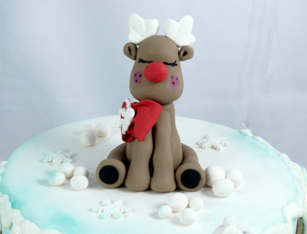 tarta cubierta con fondant y decorada con aerógrafo, abetos nevados, casitas de galleta y una figura de un reno navideño. Detalle de reno