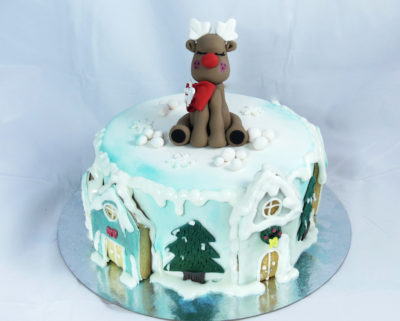 tarta cubierta con fondant y decorada con aerógrafo, abetos nevados, casitas de galleta y una figura de un reno navideño.
