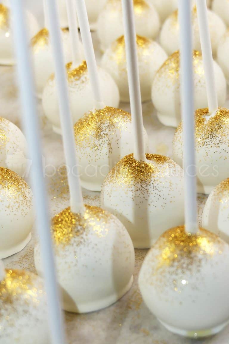 Bolita de bizcocho insertado en palo de chupa chups, bañado en chocolate blanco y decorado con purpurina comestible de color dorado.