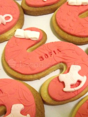Galleta de mantequilla con forma de letra "S" y detalles en fondant con forma de lazo y caballito y fondo de fondant rosa.