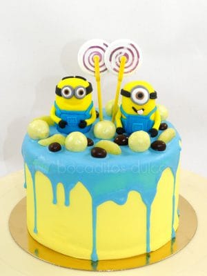 Tarta cubierta de buttercream de color amarillo con chocolate derretido por encima de color azul, decorado con chuches, y dos muñecos de la pelicula minions de fondant.