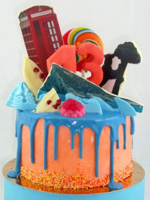 Tarta drip cake cubierta de buttercream rosa y birtas de colores, chocolate azul derretido, y decorado con diferentes tipos de dulces y diferentes temas personalizados.