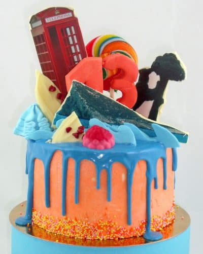Tarta drip cake cubierta de buttercream rosa y birtas de colores, chocolate azul derretido, y decorado con diferentes tipos de dulces y diferentes temas personalizados.