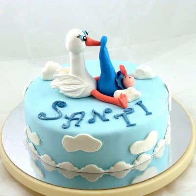 Tarta forrada de fondant azul con nubes de fondant de color blanco, nombre de niño en fondant blanco y en su base una figura de cigüeña y bebe en fondant.