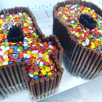 Tarta tallada en númeto cuatro, palitos de chocolate pegados con buttercream aldededor de la tarta y encima de esta lacasitos de colores, otra tarta tallada con el número cero con palitos de chocolate pegados alrededor con buttercream y decorando la superficies de esta lacasitos de colores.