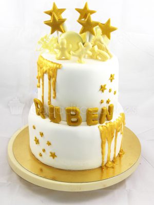 Tarta de dos pisos forrados en fondant blanco, decoracion de estrellas y nombre del niño en fondon dorado, chocolate fundido en color dorado y chocolatinas con forma de muñeco en chocolate blanco.
