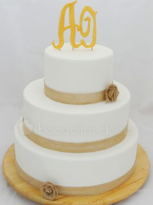 Tarta de tres pisos cubierta de fondant blanco y una cinta de rafia marron alredesor de todoas ellas, y coronada con dos letras en fondant.