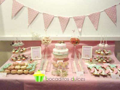 Mesa dulce decorada con tonos pastel, compuesta por Cake pops cubiertos con candy mels en tonos verde y rosa decorados con bolitas de caramelo en color blanco. Galletas de mantequilla con motivos de bebe decorados con fondant de color verde, rosa, y blanco. Tarros de cristas rellenos de chuherias, donuts con glaseado de color verde y rosa decorados con bolitas de caramelo color blanco, y tarta de dos pisos cubierta de fondant blanco el prier piso y fondant rosa el segundo, decorados los dos pisos con lunares de distintos tamaños de color rosa y verde y en la base un osito de fondant de color marron tumbado encima de una mantita.
