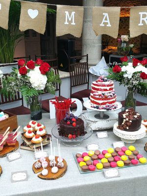 Mesa dulce en color blanco y rojo compuesta por minicupcakes de diferentes sabores, mini tartaletas de nata y fresa, cake pops en color blanco y marron, decorados con bolitas de caramelo de color rojo y otros con chocolate fundido en forma de rallas, tarta cuberta de chocolate con adornos tambien de chocolate, tarta drip cake con chocolate derretido y decorada con frutas del bosque y tarta de dos pisos desnuda decorada con frutos rojos, mini vasos, galletas de mantequilla con forma de tacon en color rojo y en forma de bigote en color negro, diferentes tipos de chuches, y para terminar deliciosos macarons de color amarillo, rosa y marron.