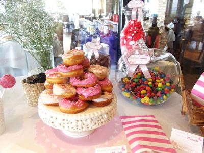 Mesa dulce de estilo vintage en colores rosos y blancos, con donuts con glaseado blanco y rosa, cakepops cubiertos con candy mels blanco y rosa y pequeños cristalitos de caramelo en color rosa, ninicupcakes con buttercream de fresa y vainilla, botellas de cristal con batido de fresa y pequeños paquetes de palomitas.