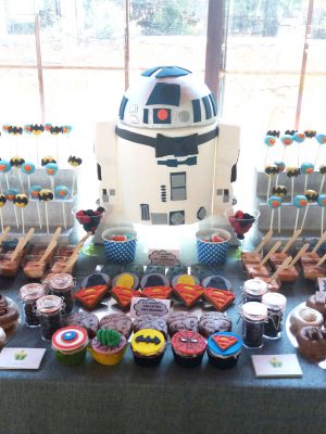 Mesa dulces compuesta por cupcakes y minicupcake de distintos sabores decorados con fondant dondo forma a diversos superheroes,cake pops cubiertos de candy mel azul y decorado con el simbolo de superman en fondant, cake pops cubierto de candy mel amarillo y decorado con el simbolo de batman en fondant, mini vasos con bizcocho, donunt con glaseado color blanco y chocolate decorados con distintos tipos de chocolatinas con formas de personajes de star wars, tarros de cristal rellensos de gominolas, galletas de mantequilla redondas decoradas con fondant distintos personajes de star wars, galletas de mantequilla con la forma del simbolor de superman decoradas con fondant, diferentes tipos de chucherias, y tartata tallada con el pesonaje de la pelicula star wars r2d.2