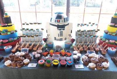 Mesa dulces compuesta por cupcakes y minicupcake de distintos sabores decorados con fondant dondo forma a diversos superheroes,cake pops cubiertos de candy mel azul y decorado con el simbolo de superman en fondant, cake pops cubierto de candy mel amarillo y decorado con el simbolo de batman en fondant, mini vasos con bizcocho, donunt con glaseado color blanco y chocolate decorados con distintos tipos de chocolatinas con formas de personajes de star wars, tarros de cristal rellensos de gominolas, galletas de mantequilla redondas decoradas con fondant distintos personajes de star wars, galletas de mantequilla con la forma del simbolor de superman decoradas con fondant, diferentes tipos de chucherias, y tartata tallada con el pesonaje de la pelicula star wars r2d.2