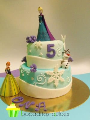 Tarta de dos pisos cubierta de fondant blanco y pintada ligeramente en color azul con tinta comestible, adornada con copos de nieve en fondant blanco y pequeñas flores moradas de fondant, el muñeco olaf en fondant y las dos princesas portagonistas en muñecas de platico.