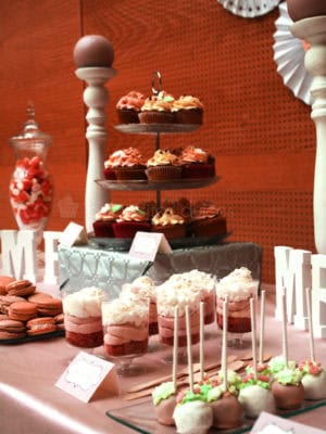 Variedad de gominolas, vasitos con vizcocho de red velvet, buttercram de fresa y nata, cupcakes de chocolate con buttercream de fresa y dulce de leche, macrons, cukepops bañados en cany mels de color blanco y rosa.