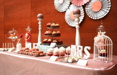 Variedad de gominolas, vasitos con vizcocho de red velvet, buttercram de fresa y nata, cupcakes de chocolate con buttercream de fresa y dulce de leche, macrons, cukepops bañados en cany mels de color blanco y rosa.