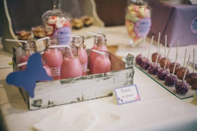 Cake pops cubiertos de candy mels de color morado y naranja decorados con bolitas blancas y moradas, botellas rellenas de batido de fresa y variedad de gominolas