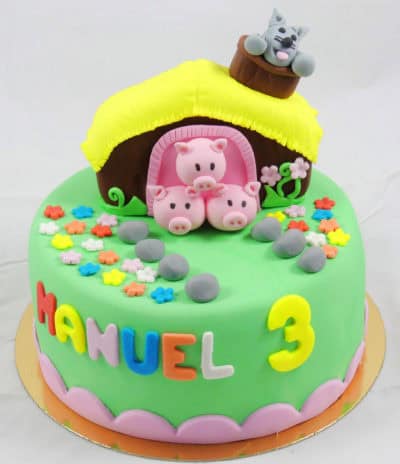 Tarta cubierta de fondant de color verde, con el nombre de Manuel en diferentes colores de fondant, el numero tres en fondant amarillo, folres de diferentes colores en fondant, los tes cerditos y el lobo en una pequeña casa en fondant y pequeñas piedras tambien en fondant.