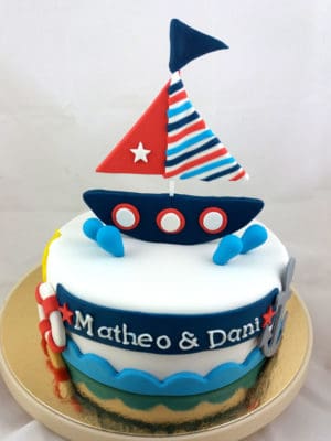 Tarta cubierta de fondant blanco, con una pequeña anacla de fondant, y flotador tambien en fondant, el nombre de Matheo y Dani en fondant blanco, pequeñas gotas de fondant azul y un gracios barco en fondant sobre la tarta.