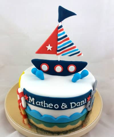 Tarta cubierta de fondant blanco, con una pequeña anacla de fondant, y flotador tambien en fondant, el nombre de Matheo y Dani en fondant blanco, pequeñas gotas de fondant azul y un gracios barco en fondant sobre la tarta.