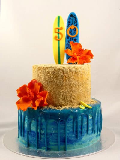 Trata de dos pisos, el de la base cubierto de buttercream azul y ganache chorreando alrededor, y la segunda cubierta de buttercream con galleta y decorada con dos tablas de surf de fondant y flores de fondant color naranja.