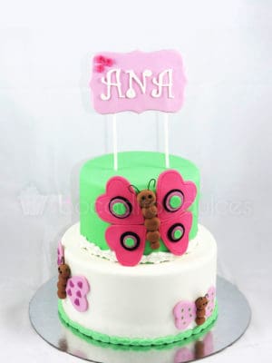 Tarta de dos pisos forrada de fondat blanco y la mas pequeña de fondnat verde, pequeñas mariposas de fondant, y otra mariposa mas grande en fondant, con el nombre de Ana en un letrero tambien de fondant