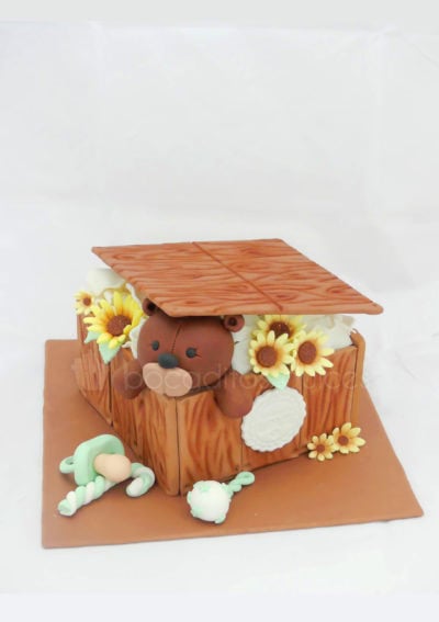 Tarta cuadrada forrada de fondant simulando tablas de madera, pequeñas flores amarillas de fondant, pequeño sonajero de fondant, chupete tambien de fondant, y osito tambien de fondant