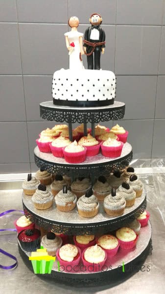Torre de dulces compuesta por una tarta forrada en fondant blanco, con pequeñas bolitas negras de fondant alrededor de la tarta, con dos muñecos vestidos de novios coronando la tarta, cupcakes de diferentes sabores.