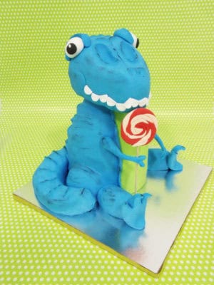 Tarta tallada con la forma de un dinosaurio, y despues forradas con fondant de color azul, y detalles en fondant como los ojos, dientes, y una pequeña piruleta.