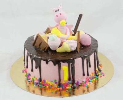 Tarta cubierta con buttercream de color rosa, pequeñas virutas de caramelo alrededor el numero uno en color amarillo, ganache de chocolate por encima y cayendo por los lados diferentes tipos de chocolates y gominolas sobre la tarta junto con un pequeño dragon de fondant